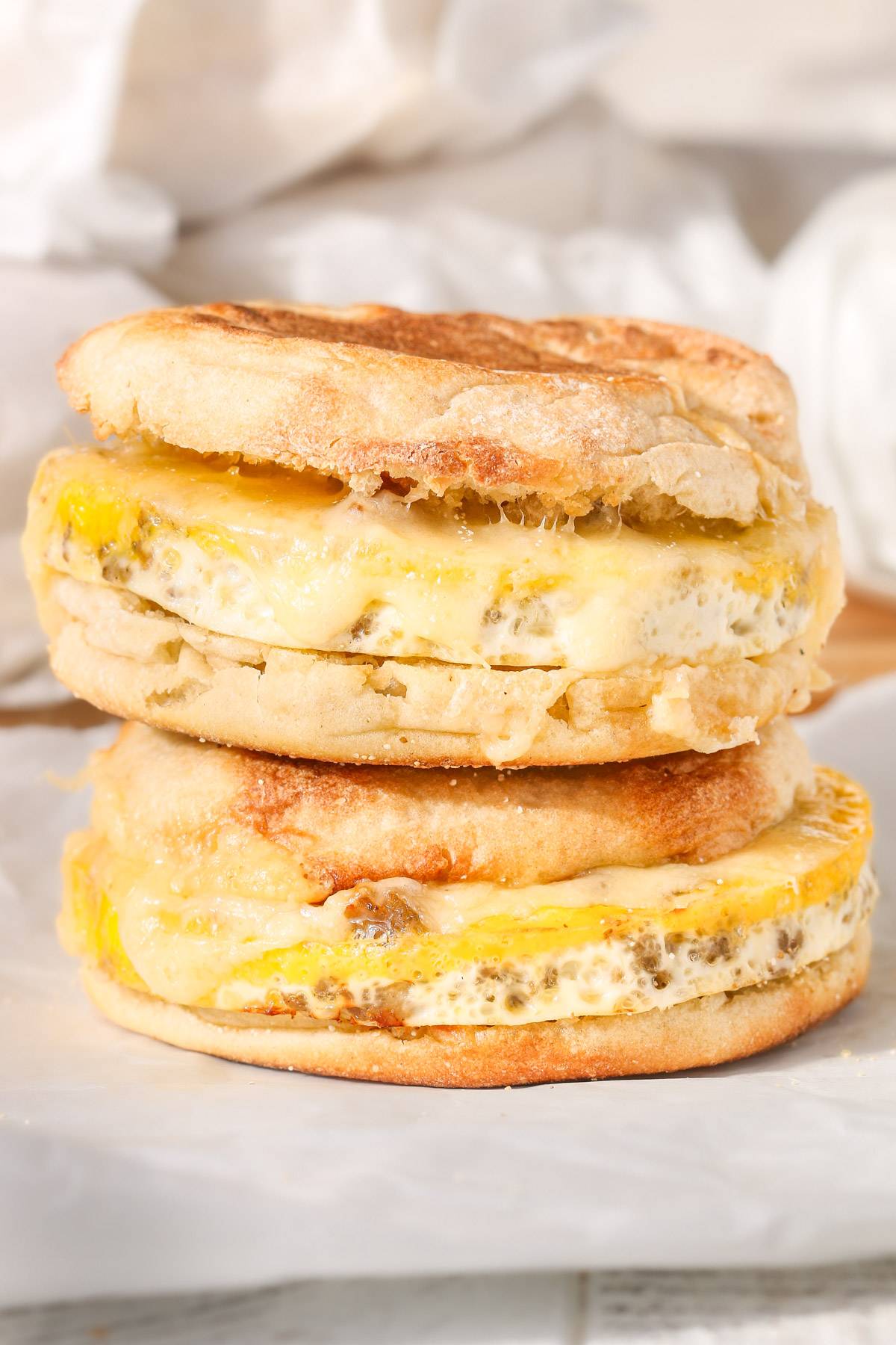 Close-up of two homemade freezer breakfast sandwiches stacked on parchment paper, made with toasted English muffins, baked egg and sausage rounds, and melted cheese.