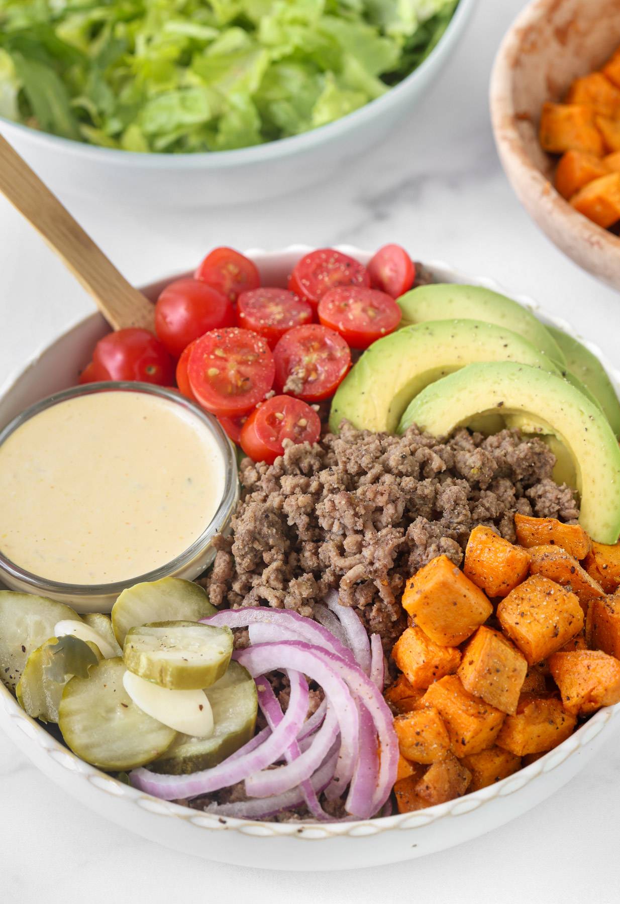 Burger bowl with seasoned ground beef, roasted sweet potatoes, avocado, cherry tomatoes, pickles, red onion, and creamy burger sauce