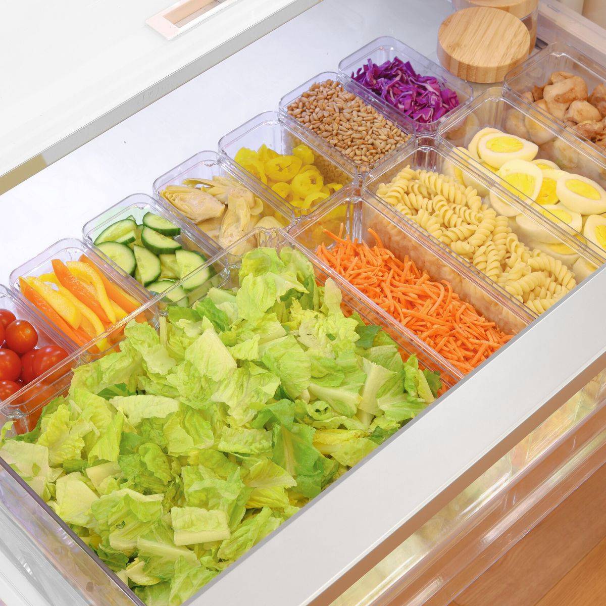 Fridge drawer salad bar with chopped romaine, cucumbers, carrots, pasta, and toppings in clear containers