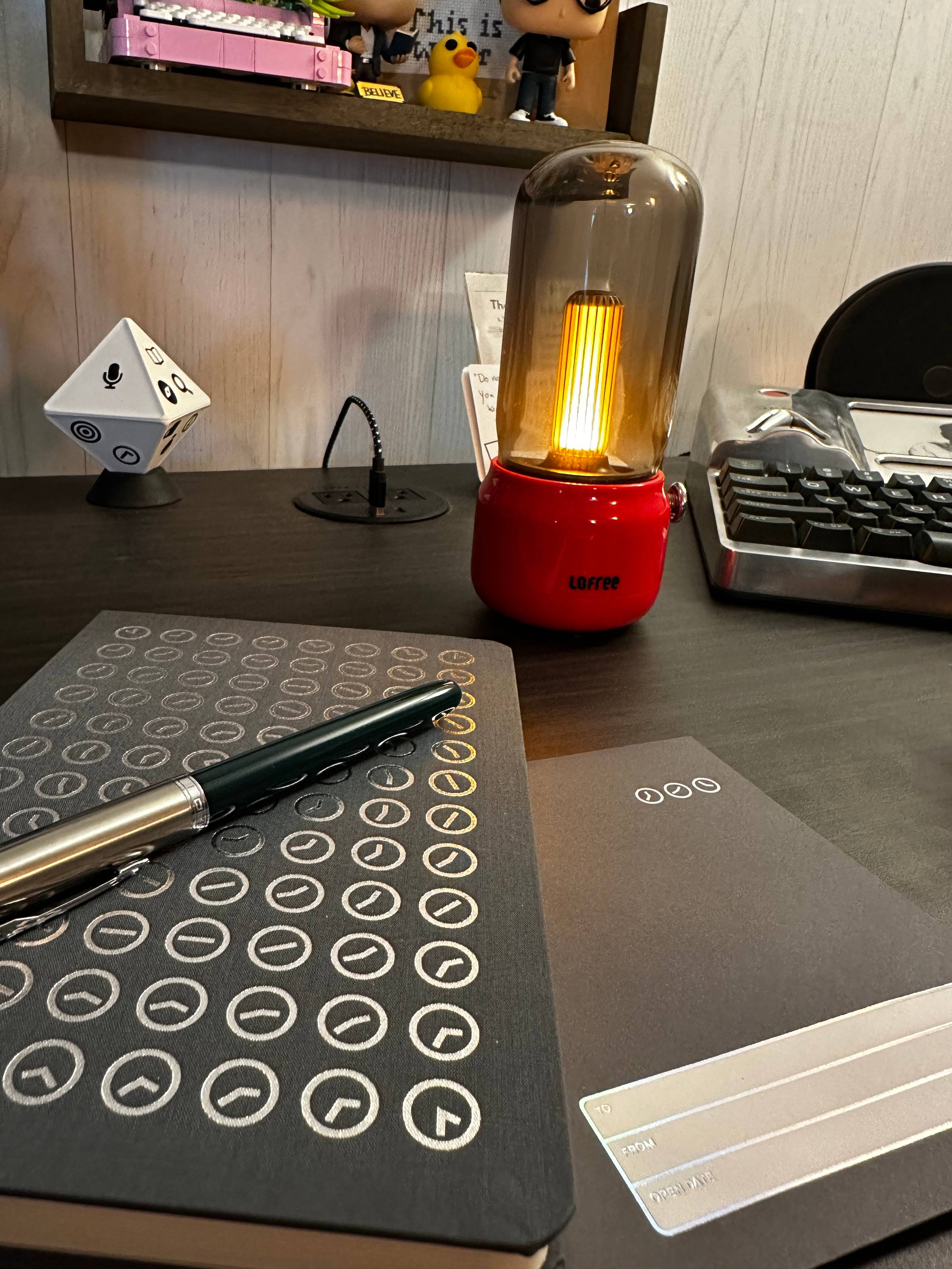 A notebook with clockcs on it and a fountain pen with a small red desk lamp behind it.