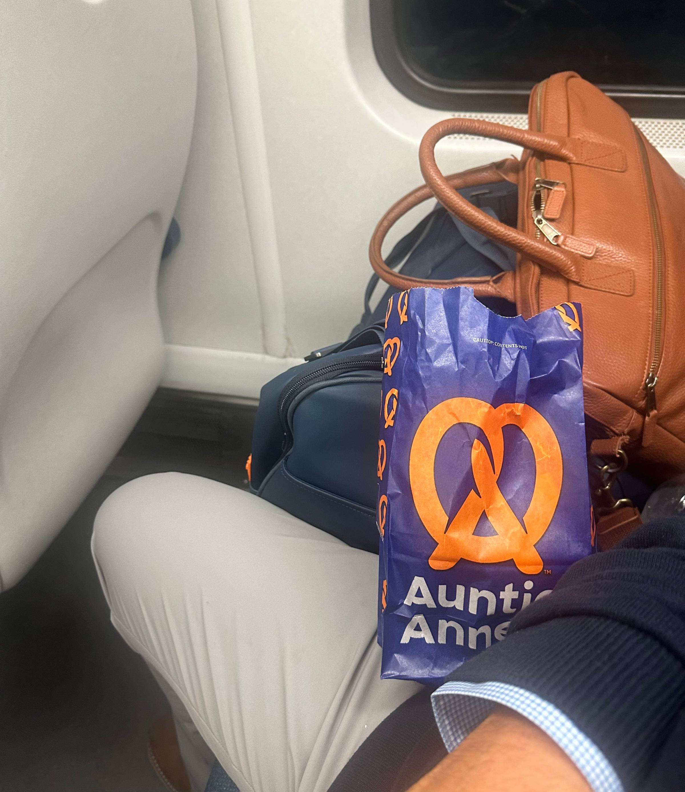 A snapshot taken on the train: Dan holding an Auntie Anne's pretzel bag, with a leather briefcase and luggage resting against the adjacent seat