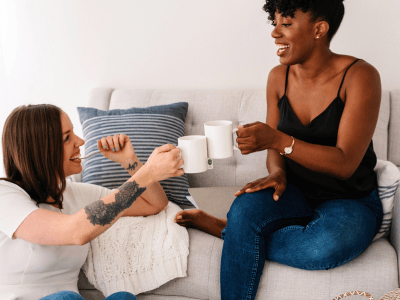 Two women sitting casually on a couch, smiling and clinking coffee mugs—natural, introvert-friendly connection in a relaxed setting.