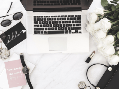 Flatlay of a laptop on a marble desk with white roses, sunglasses, planner, and feminine accessories. Keywords: workspace, lifestyle, entrepreneur.