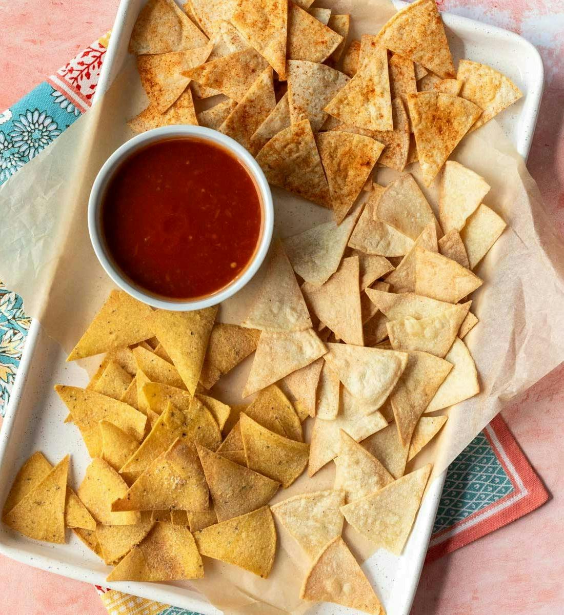 Baked and air fried tortilla chips spread out on a tray next to a cup of salsa.