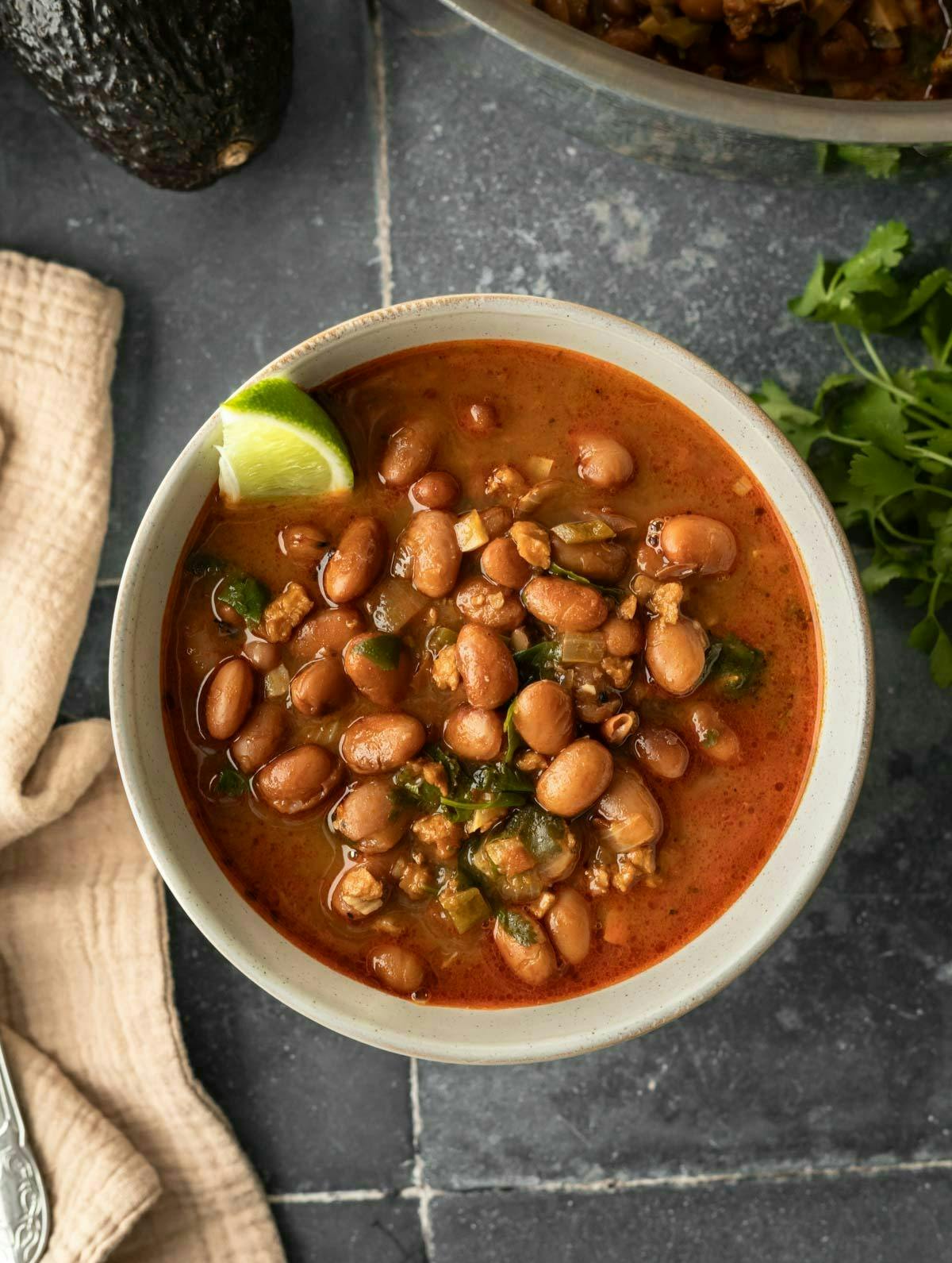 A bowl filled with charro beans with avocado and cilantro nearby.