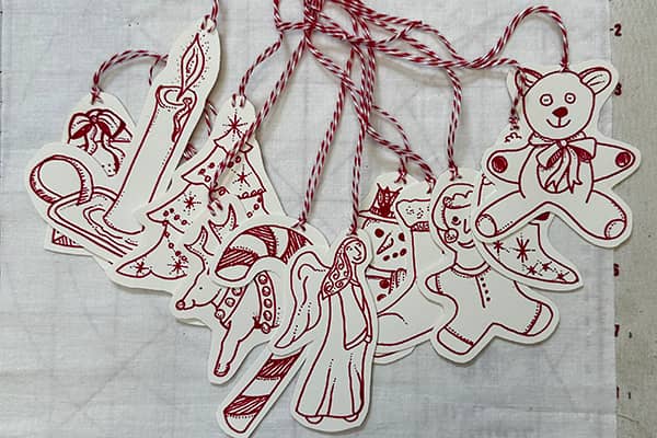 Embroidery designs were traced on hot press watercolor paper using a red pen, cut out and red & white striped cord was attached to hang each ornament with.