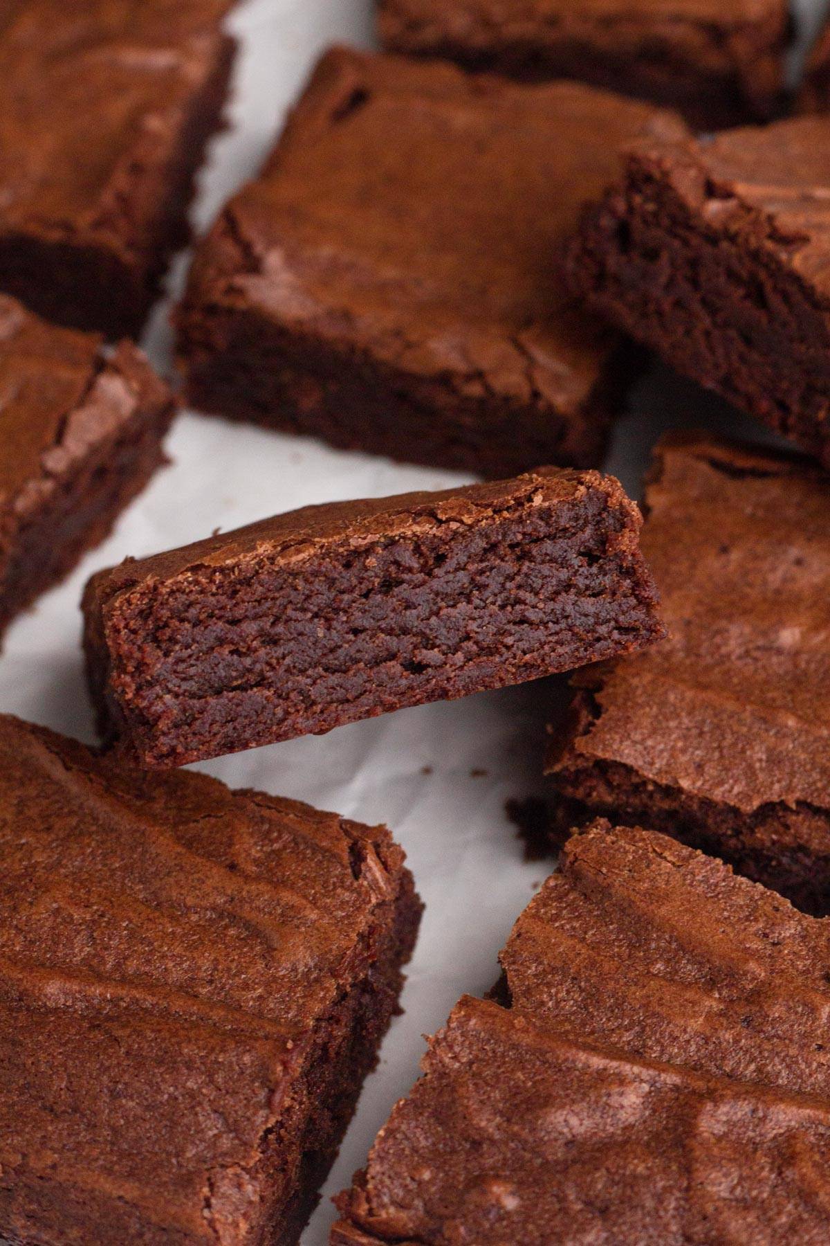 Close-up of several square-shaped chocolate brownies arranged on a light surface. One brownie is slightly lifted, showcasing its moist and dense interior. The tops of the brownies appear slightly crispy.