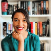 Image of Olivia Bedford sitting in front of a bookshelf with all of your favorite fantasy books. She is wearing a teal shirt and bright red lipstick and smiling. Her chin is wresting on her hand.