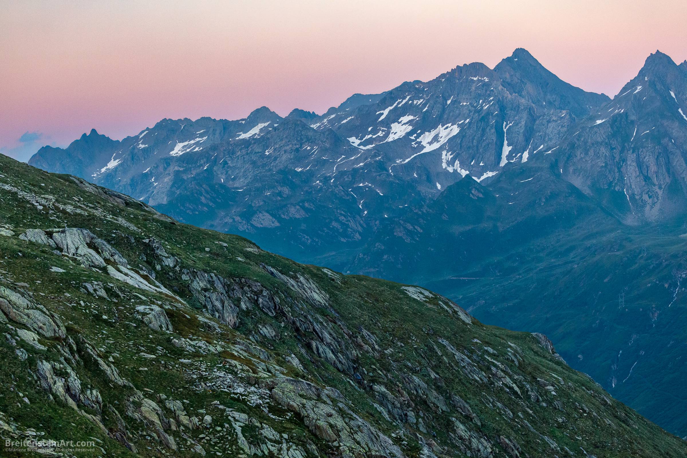 Two mountain ridges. The nearest is gray-green, the farthest is in shades of blue, with snow. The sky is pink.