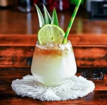 A tropical layered cocktail sits on a white fringe coaster on a wooden bar with liquor bottles in the background.