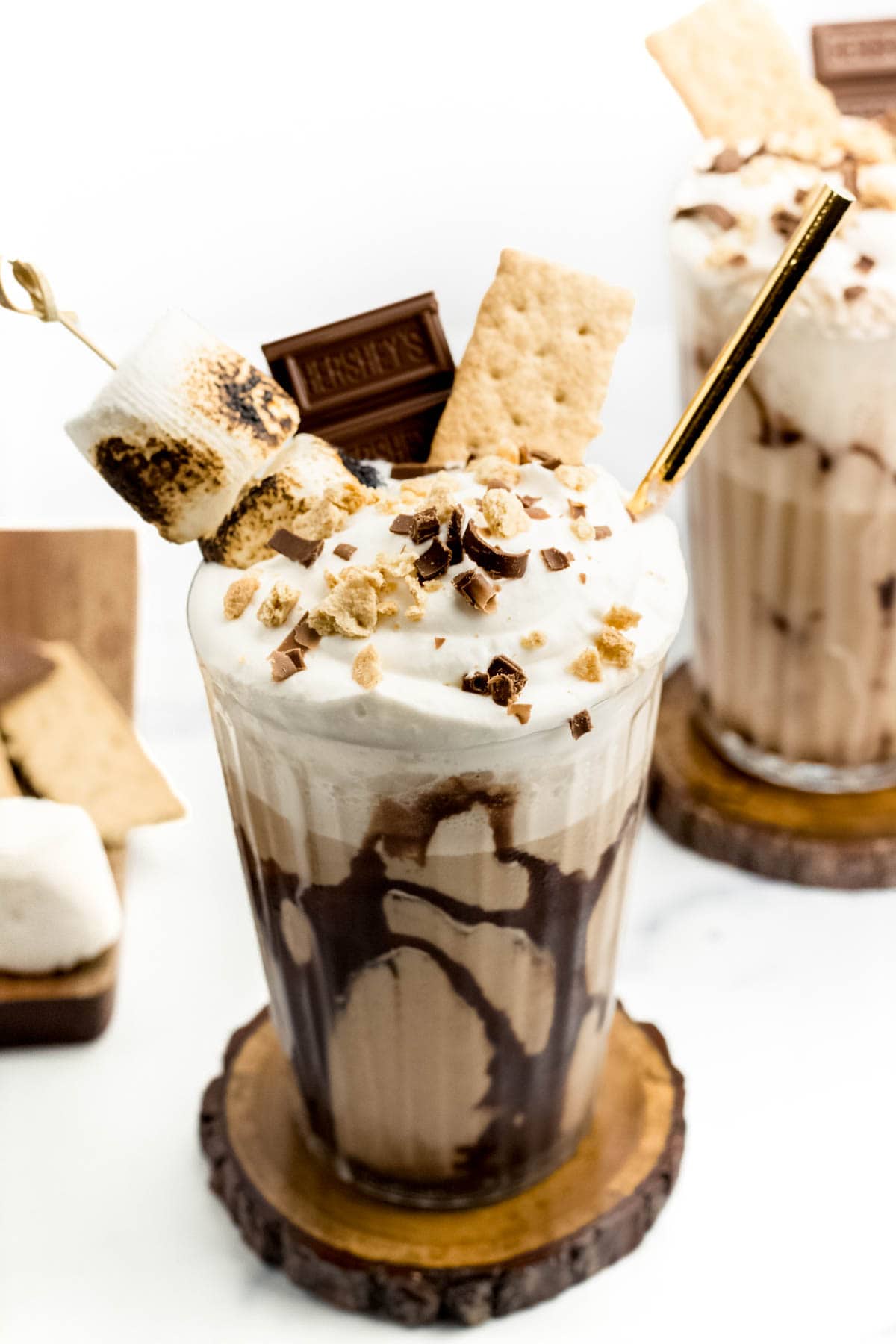 A creamy chocolate drink topped with whipped cream, graham crackers and marshmallows with a gold straw on a wooden coaster.