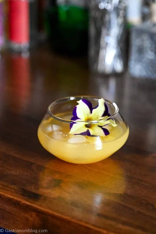 A yellow drink in a glass sitting on a wooden surface topped with a yellow and purple flower.