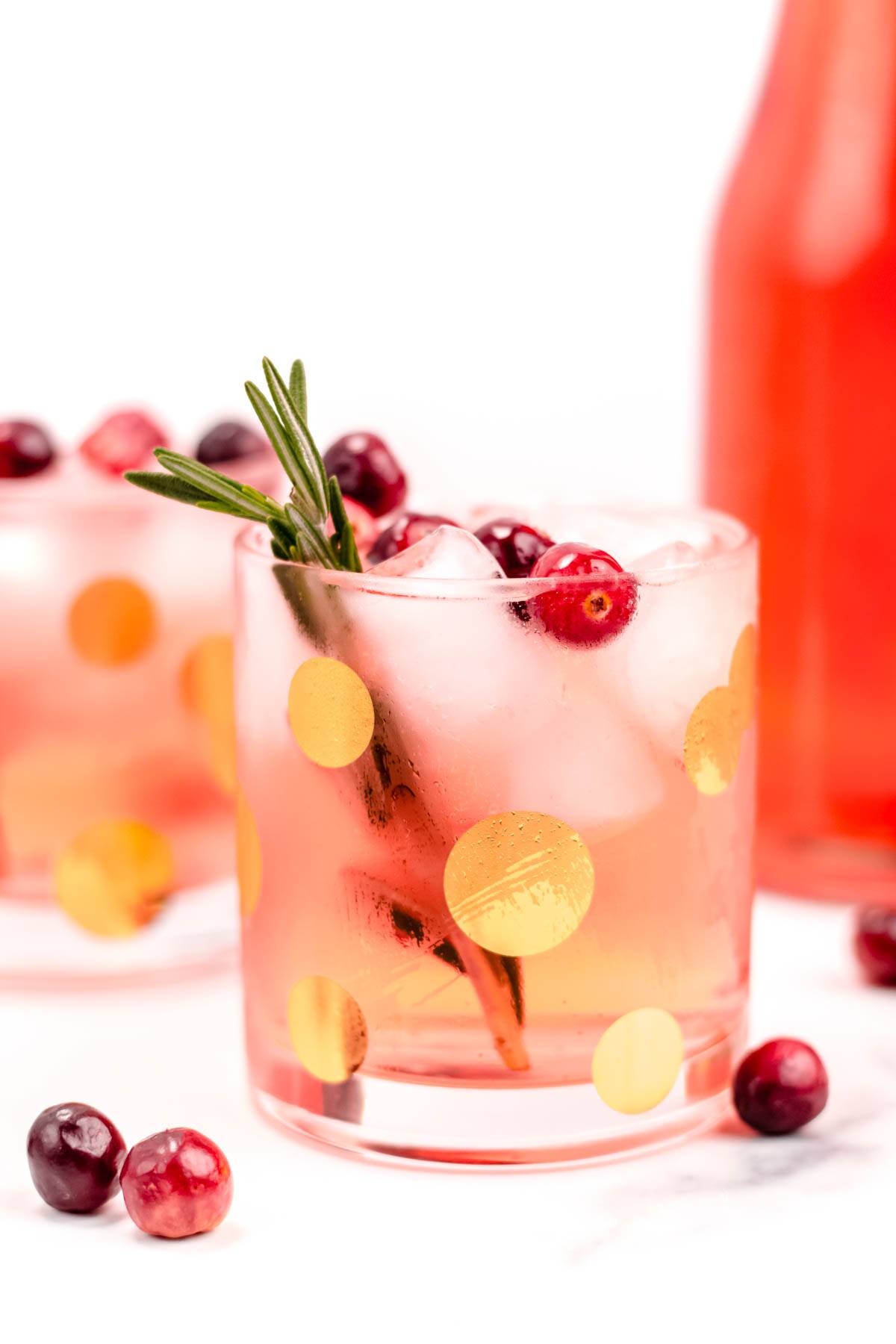 A pink drink in a rocks glass with gold polka dots garnished with a sprig of rosemary and whole cranberries.