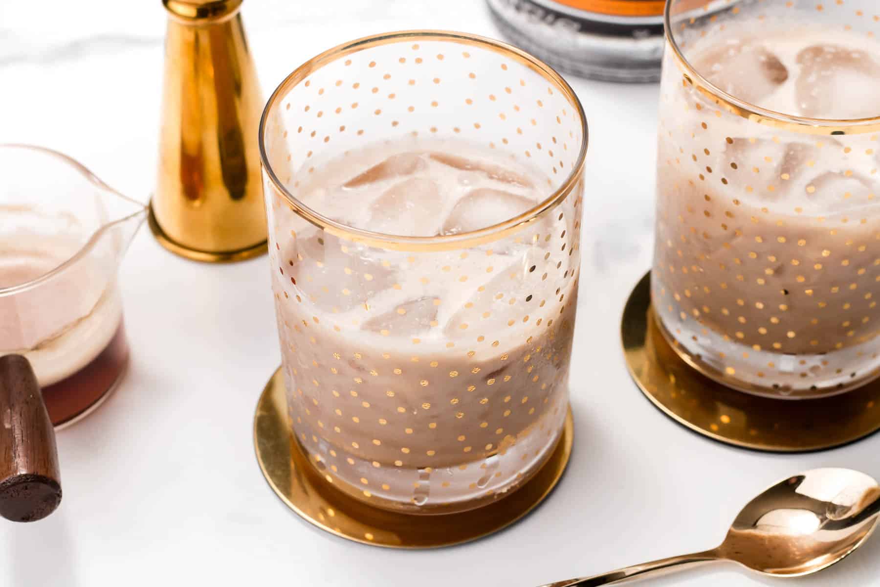 Clear rocks glasses with gold polka dots hold an iced, creamy cocktail near gold bar tools.