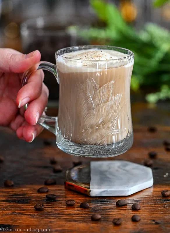 A hand lifts a glass mug filled with creamy coffee.