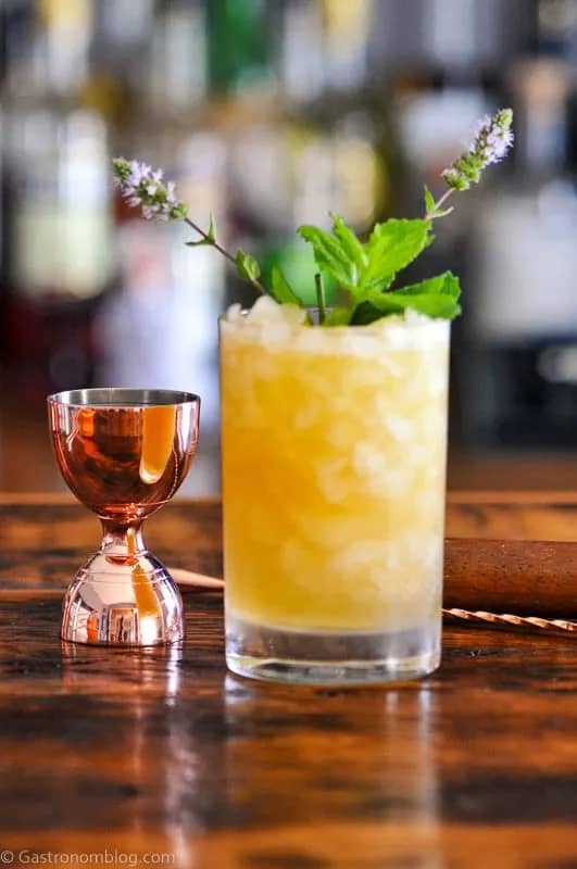 A yellow drink with crushed ice topped with herb sprigs near a copper jigger on a wooden surface.