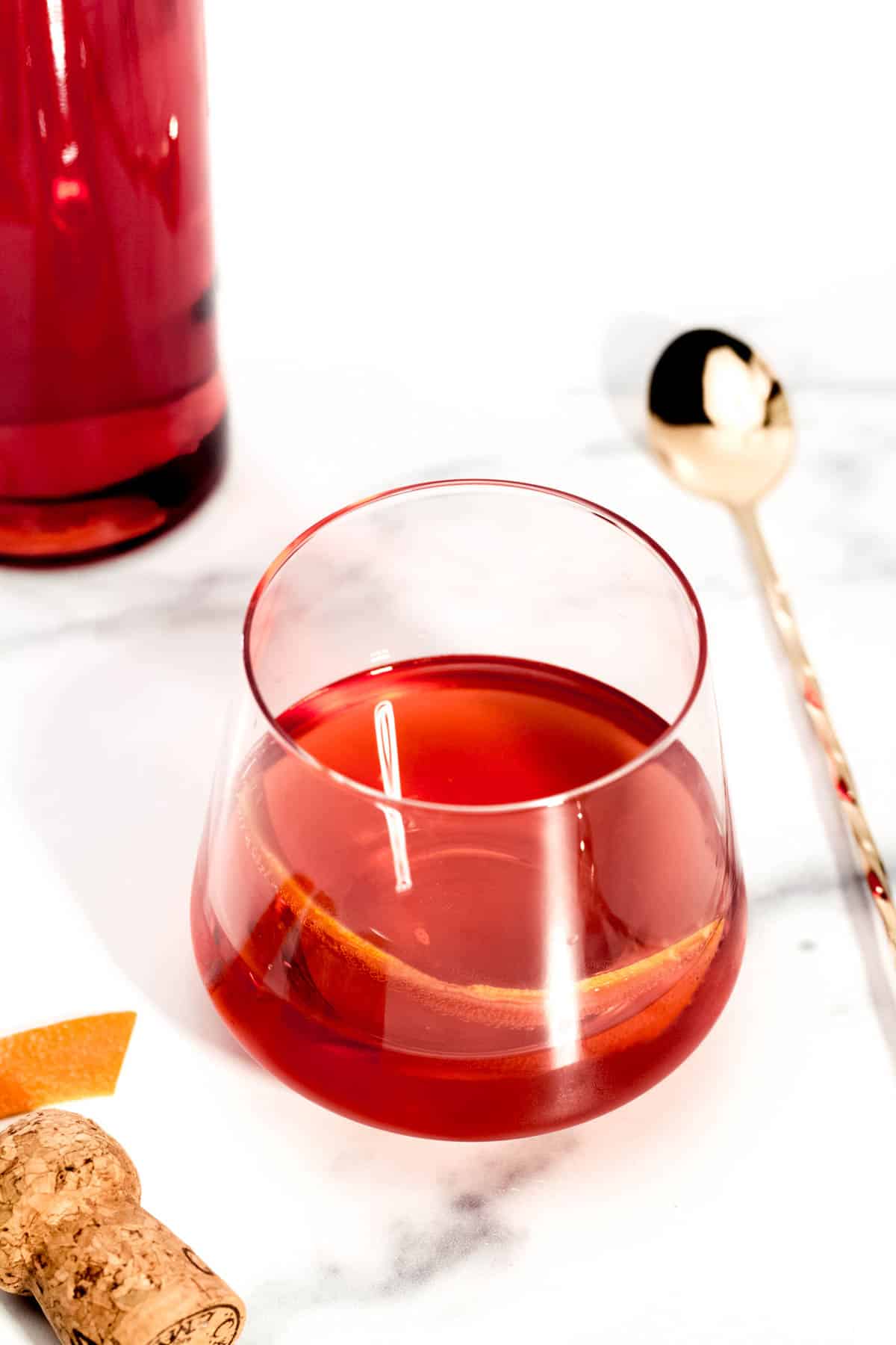 A red cocktail in a stemless glass near a cork and a gold cocktail spoon.