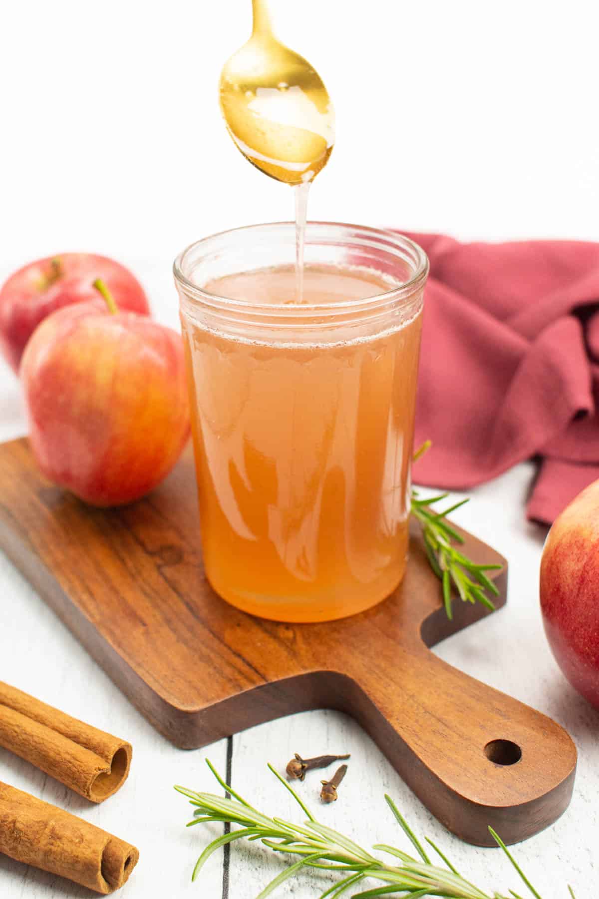 A jar of light orange syrup on a wooden cutting board with apples.