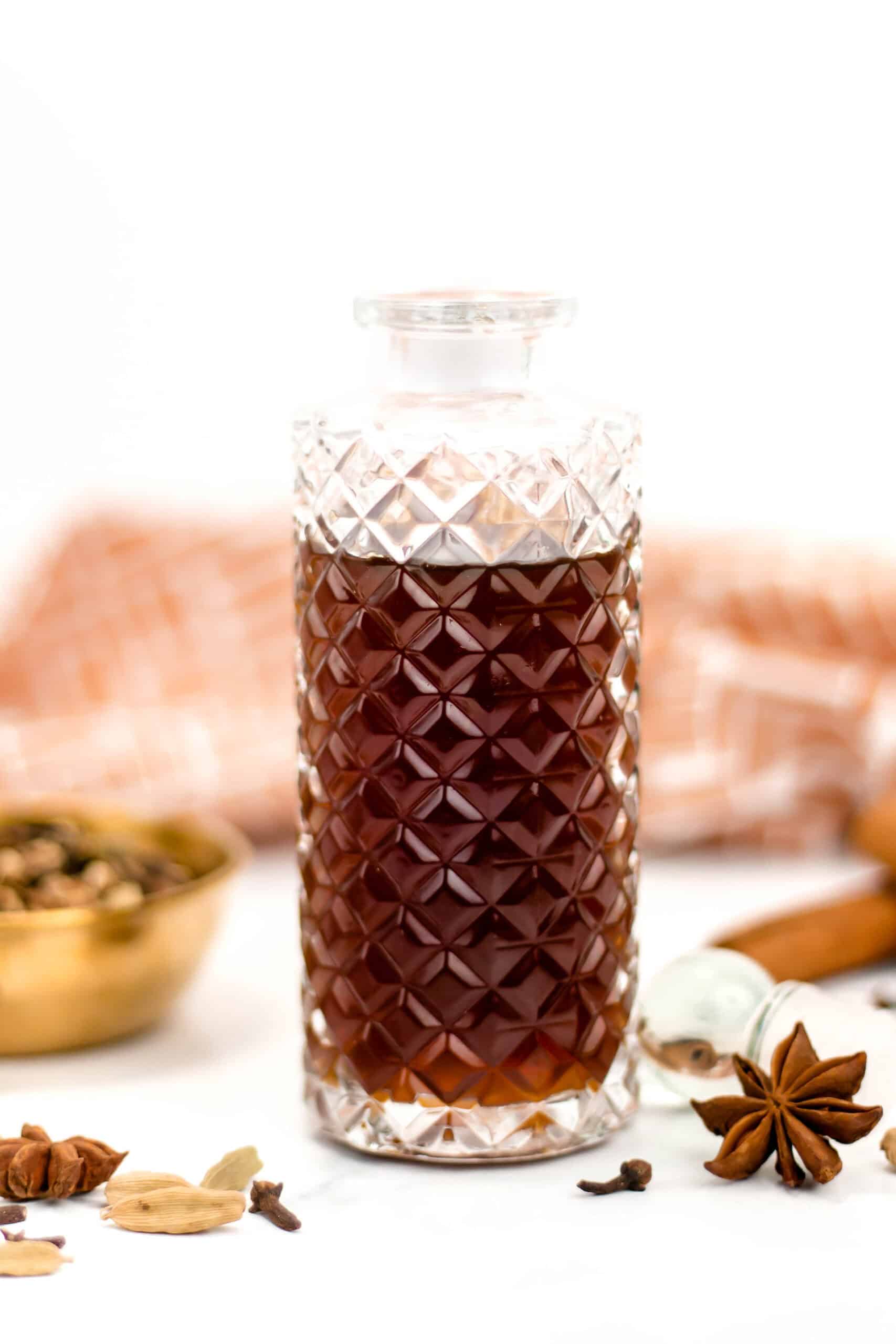 A decorative glass bottle of brown simple syrup surrounded by whole spices on a white surface.