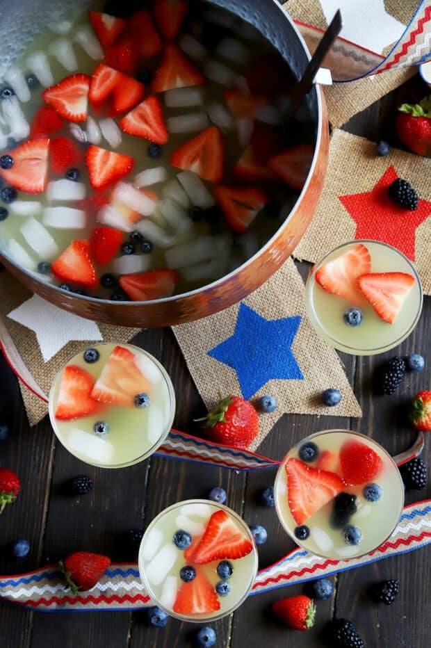 Overhead view of a copper punch bowl and glasses of pale yellow punch topped with strawberry slices and whole blueberries.