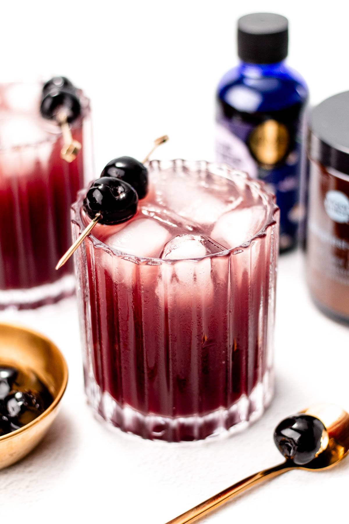 A deep red drink with ice has a gold cocktail pick holding two cherries.