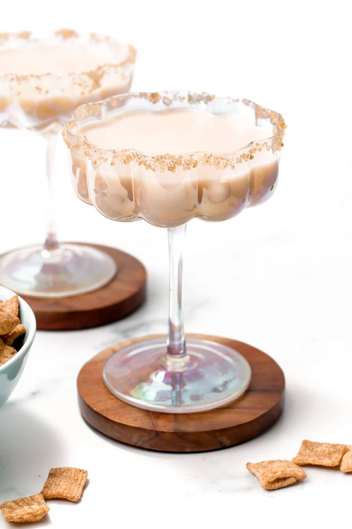 A creamy white drink in a stemmed glass sits on a round wooden coaster with pieces of cinnamon toast crunch cereal scattered around.