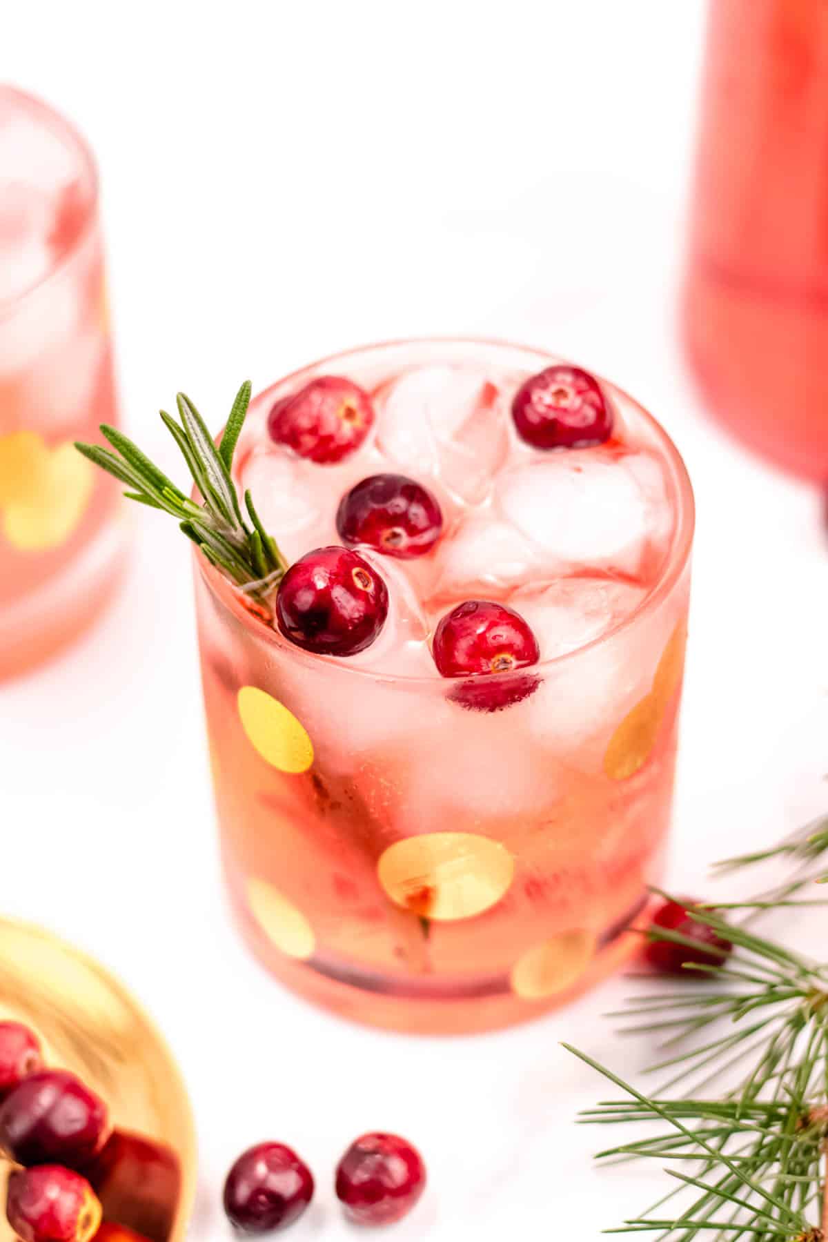 A pink cocktail with ice, whole cranberries and a sprig of rosemary.