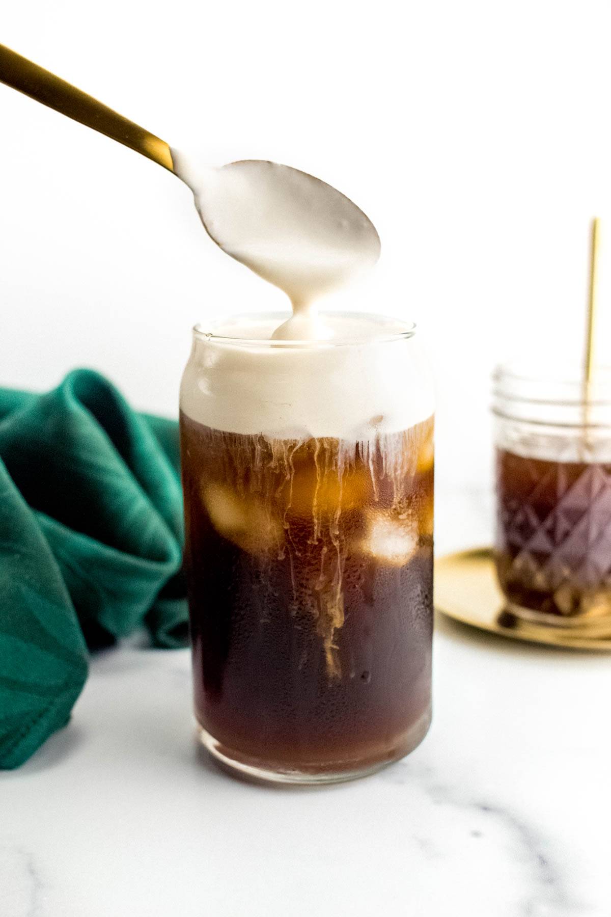 A spoon pours foam on top of a glass cup of iced coffee.