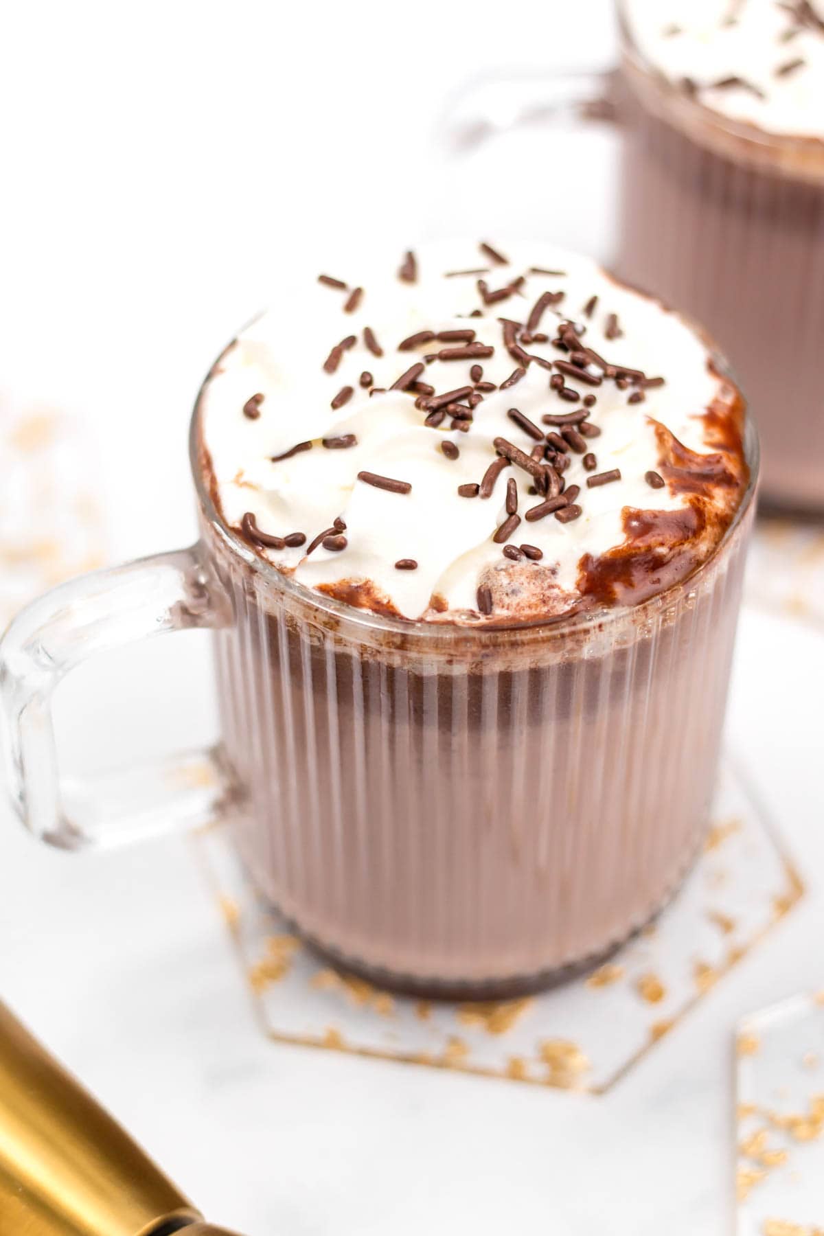 A glass mug holds a chocolate drink topped with whipped cream and chocolate sprinkles.