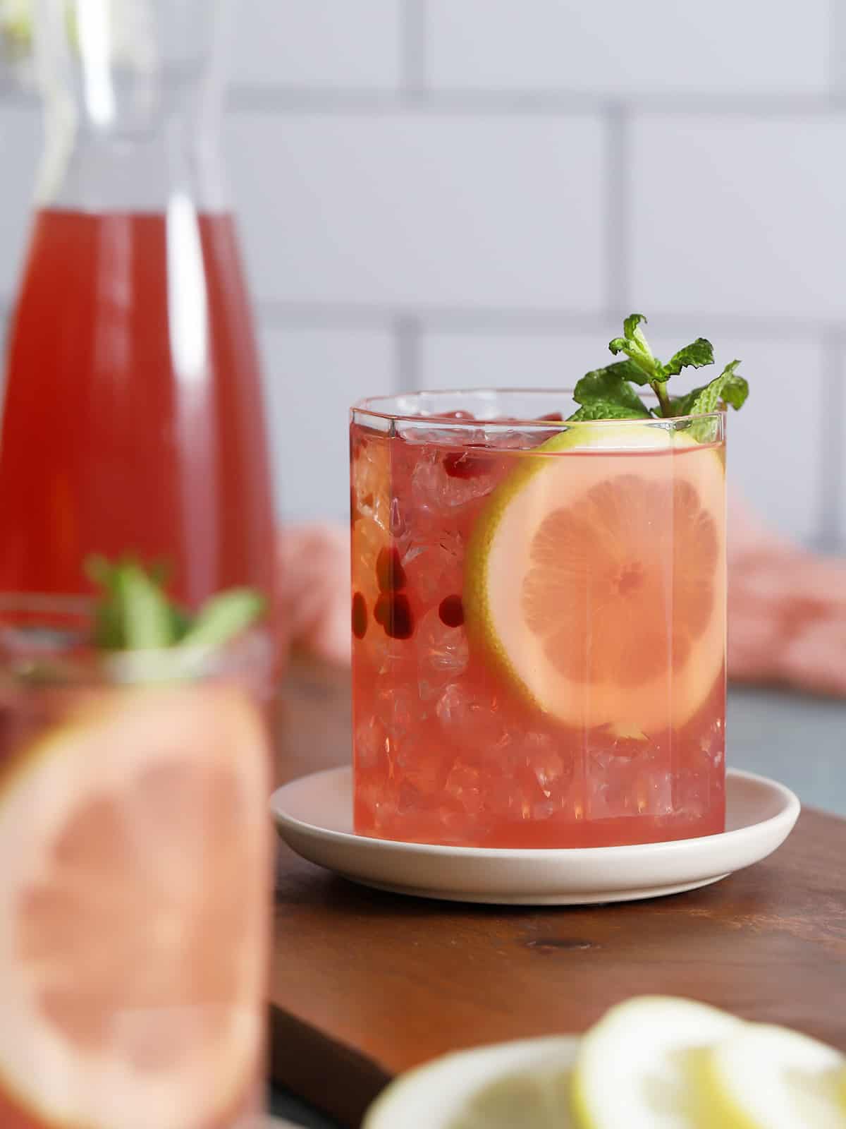 A short glass on a white plate holding a pink drink with pomagrante seeds, a lemon round and an herb sprig.