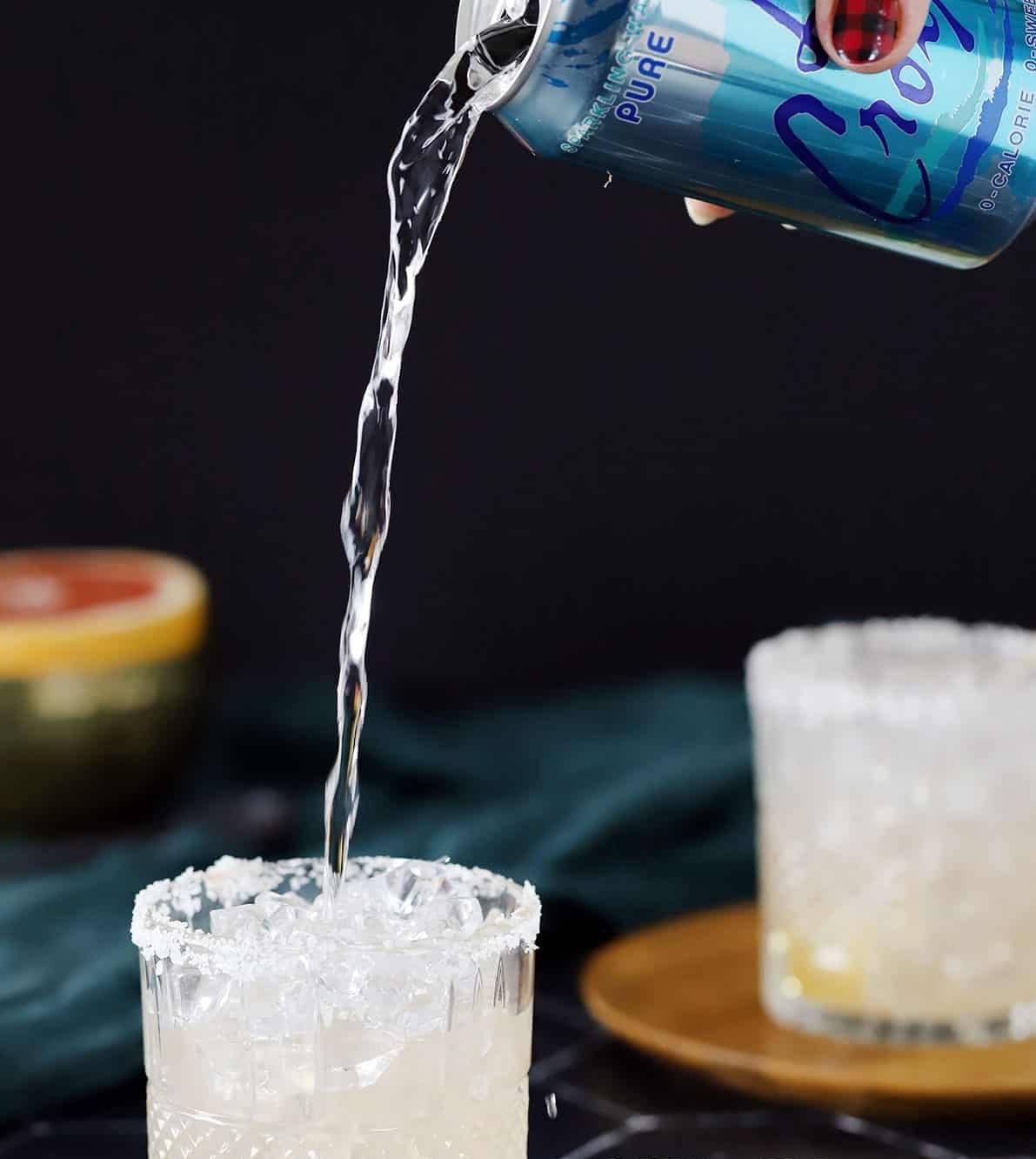 Sparkling water is poured from a blue can into a glass rimmed with salt.