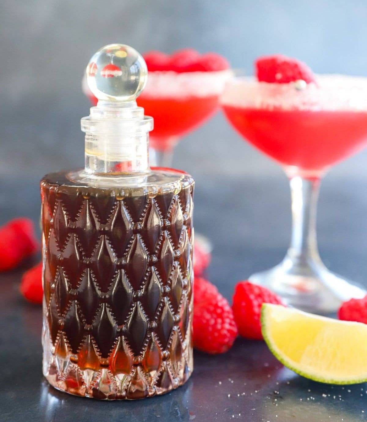 A decorative glass container of dark brown syrup near two red drinks.