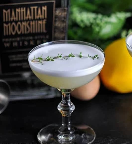 A pale drink in a stemmed glass is topped with an herb sprig.