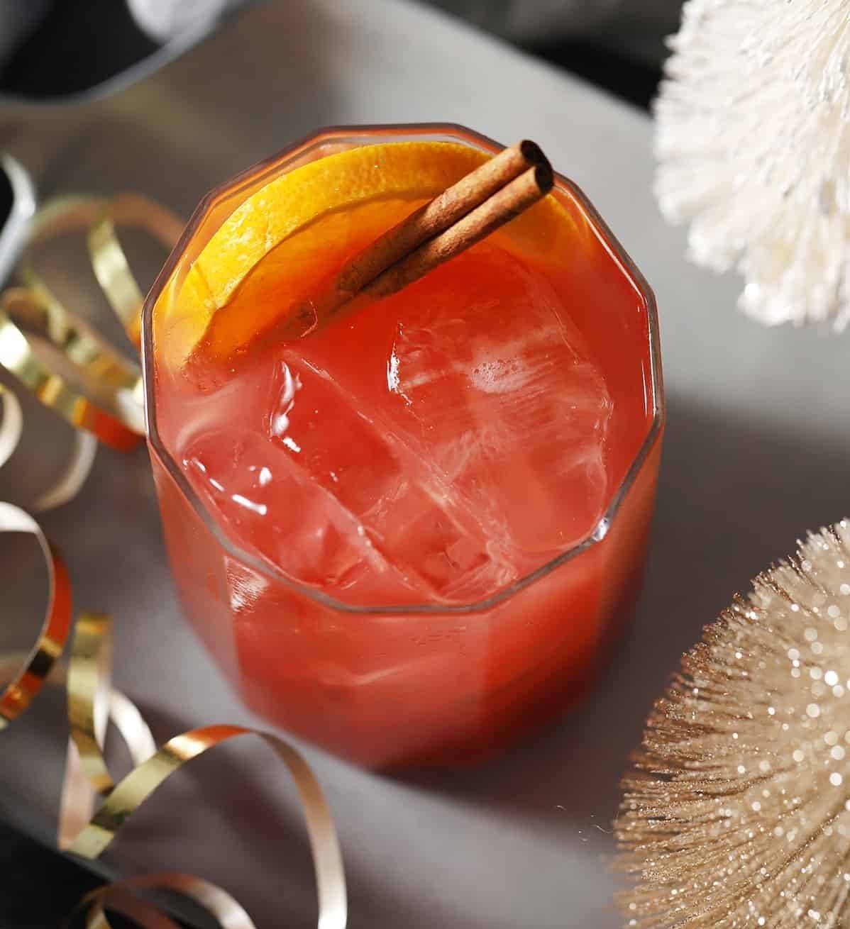 A red drink sits in a glass garnished with a cinnamon stick and a round of fruit.
