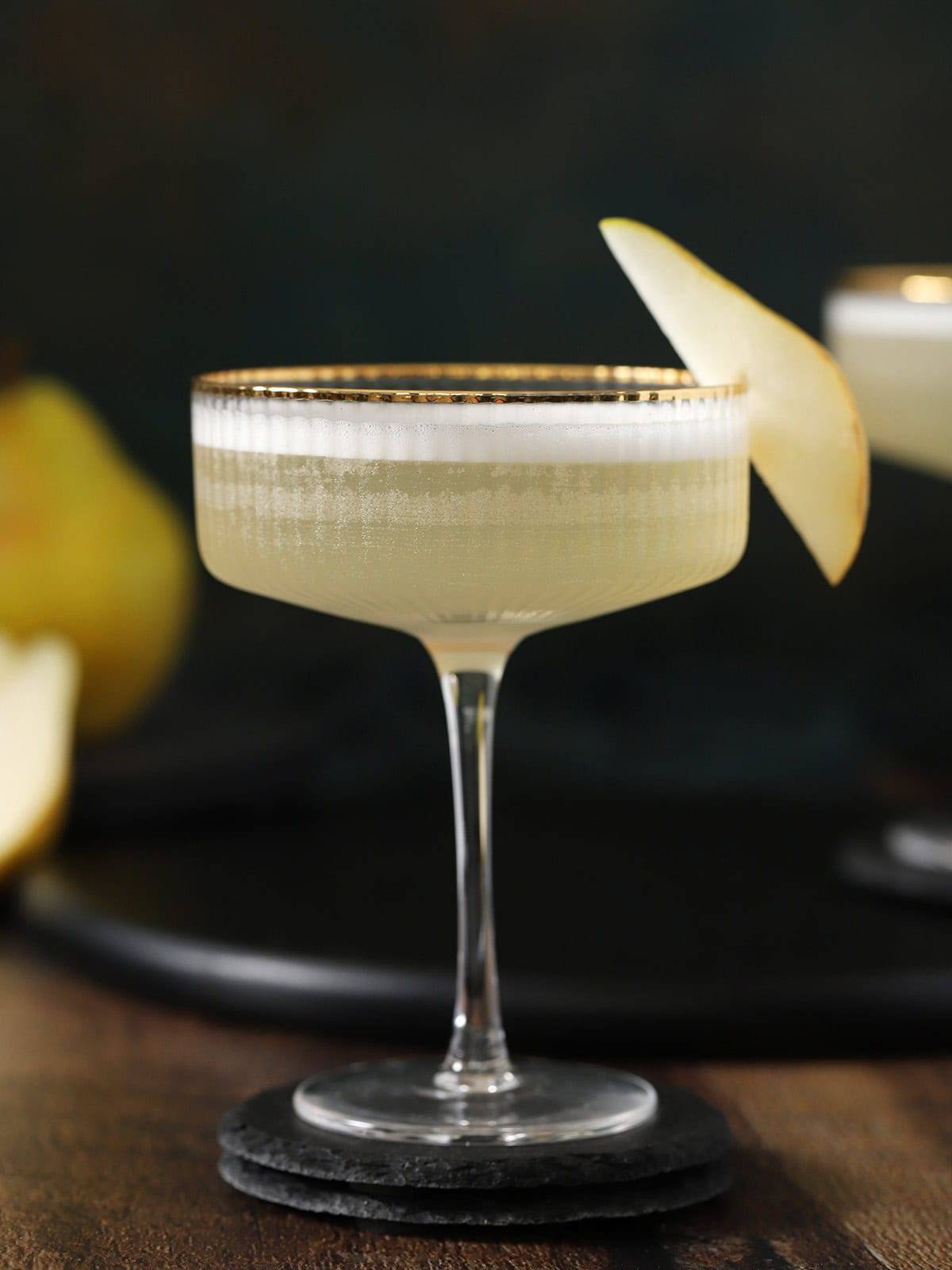 A stemmed glass holds a pale yellow cocktail with a pear slice on the edge.