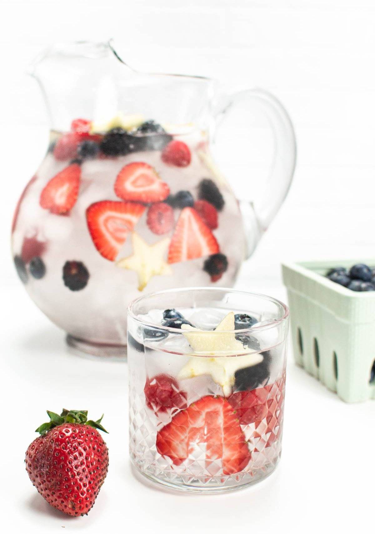 A pitcher of berry water near a glass of the water with berries and ice.