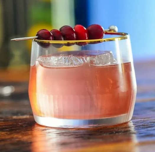 A pink drink in a glass with a cocktail skewer of cranberries sitting across the top.