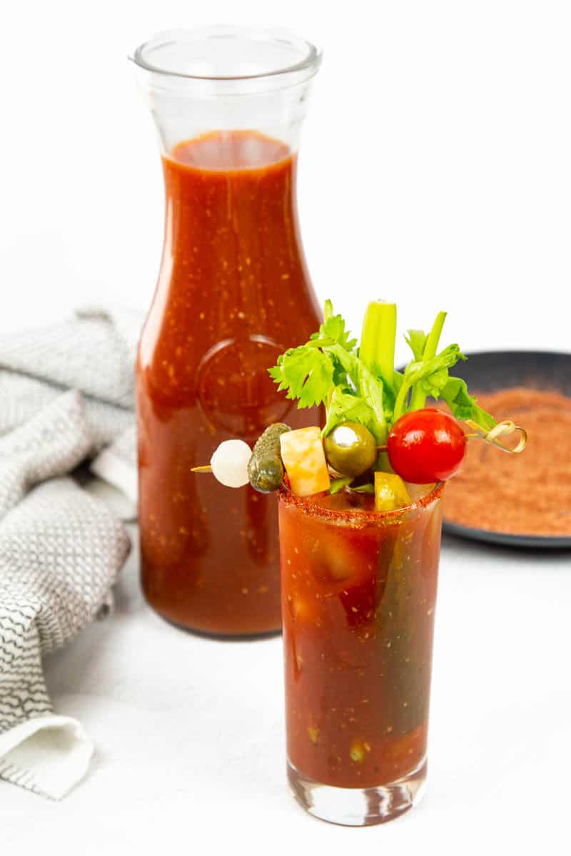 A bloody mary garnished with veggies in front of a tomato mixture in a pitcher.