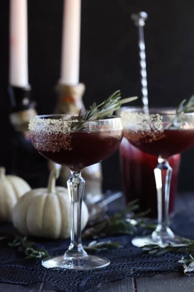 Deep purple drinks in stemmed glasses sit near small white pumpkins with a candlestick in the background.