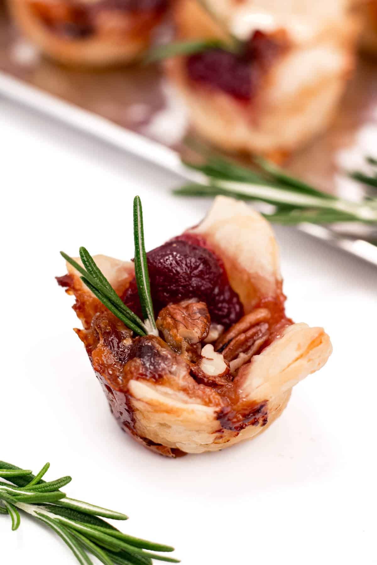 A puff pastry bite filled with cranberry and brie in front of a silver tray near rosemary garnish.
