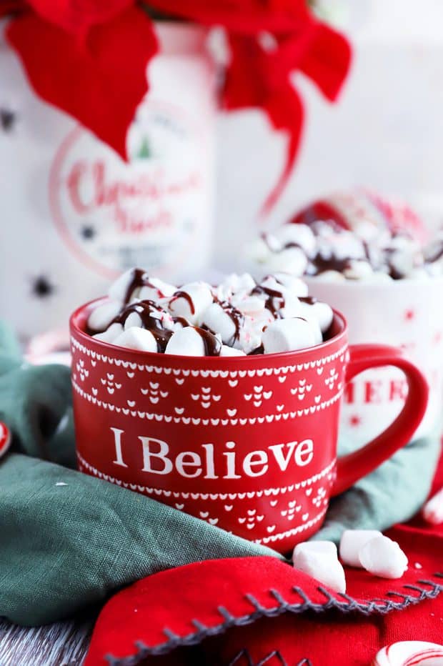 A red mug that reads 'I believe' holds a mountain of marshmallows drizzled with chocolate sauce and sprinkles.
