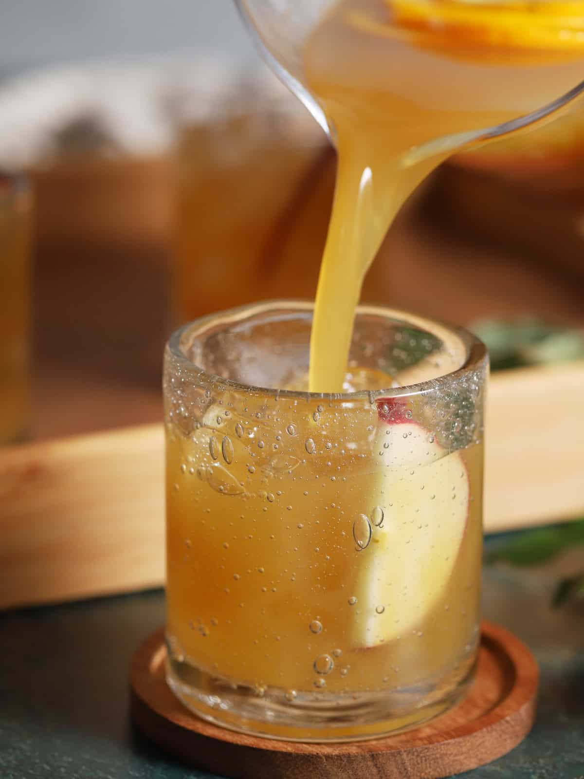 An amber liquid is poured from a glass pitcher into a short glass with an apple slice.