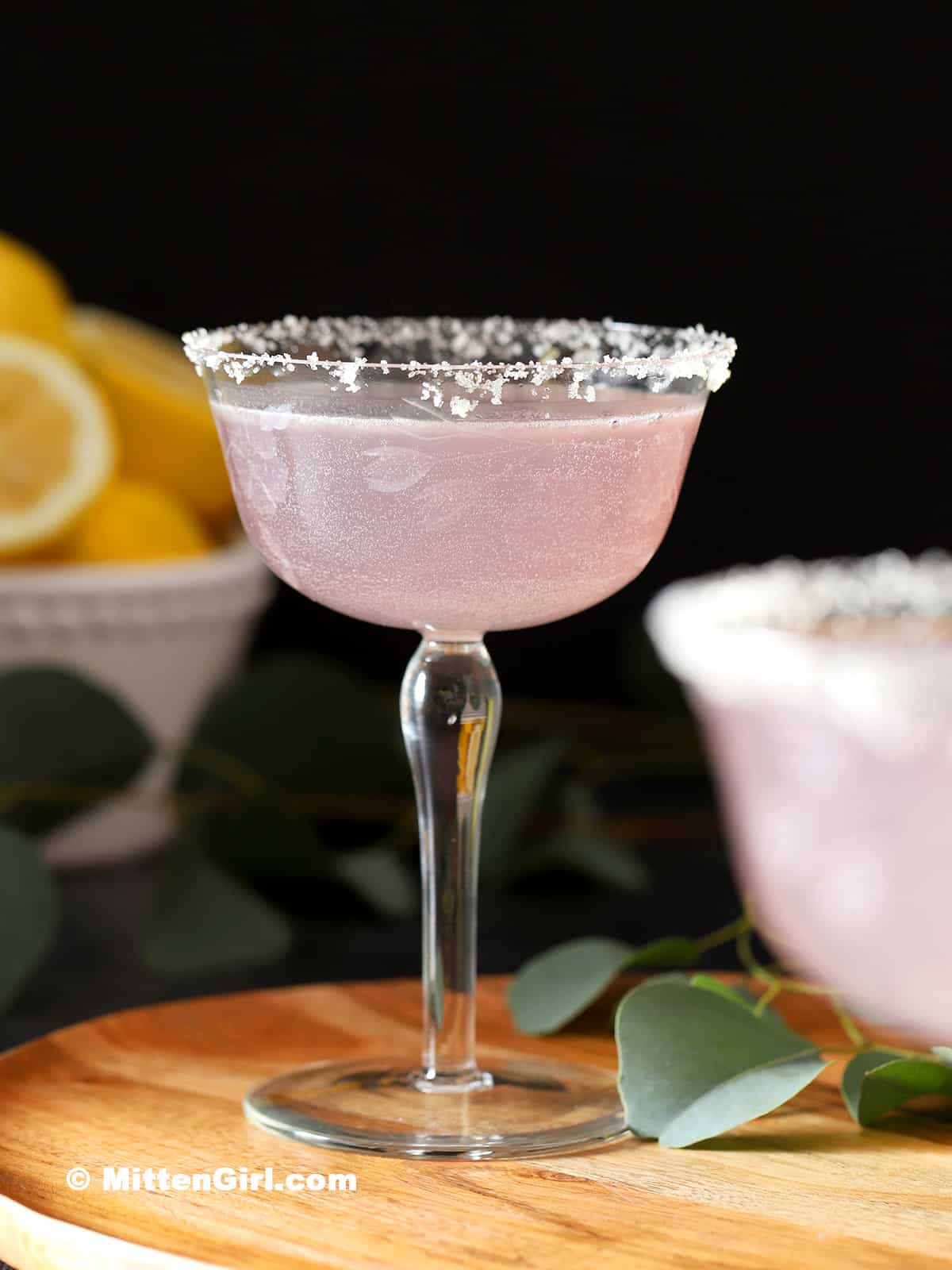 A stemmed glass holds a pale purple drink rimmed with sugar.
