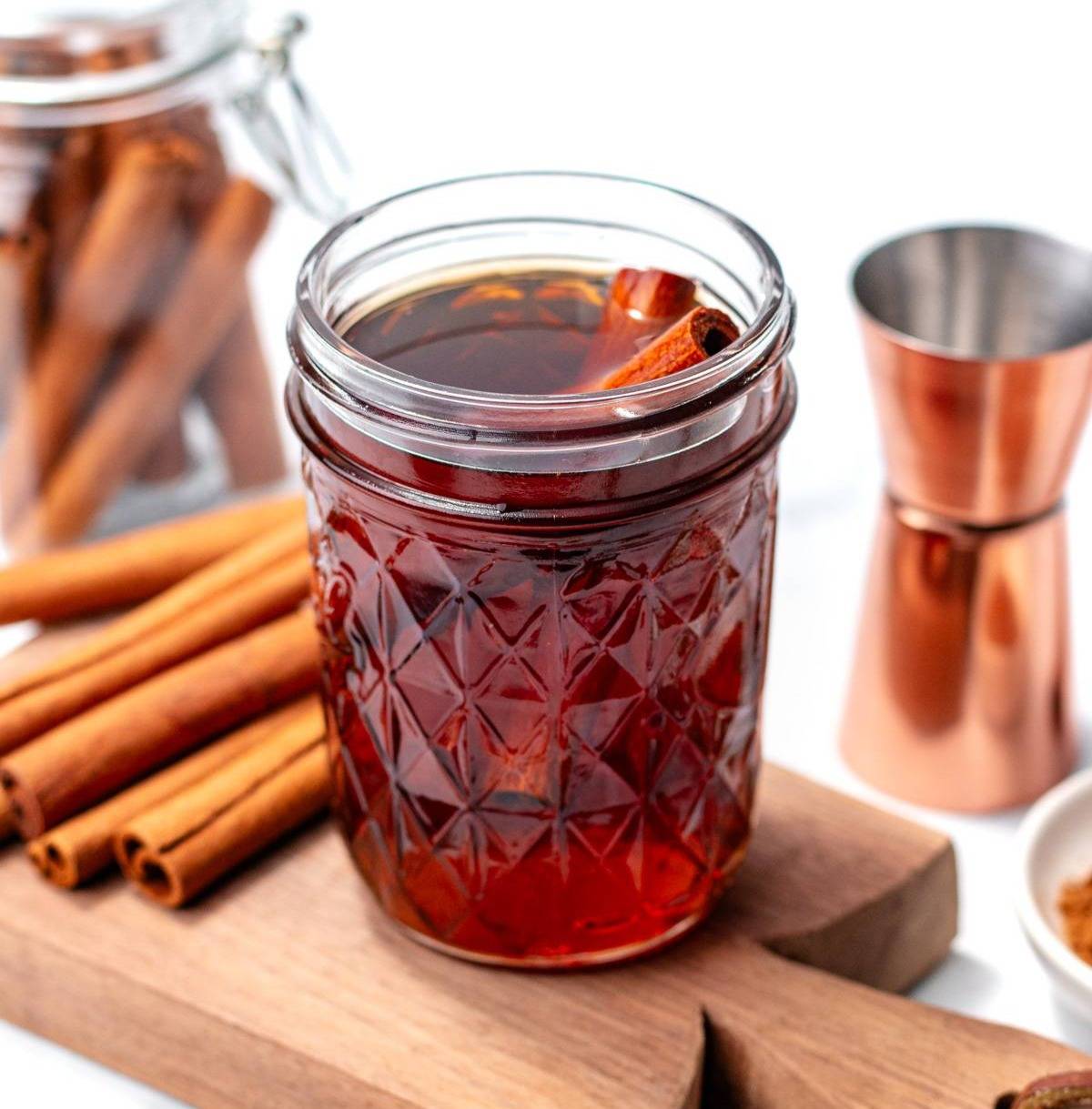 Cinnamon syrup with cinnamon sticks in a jar next to more cinnamon sticks, a copper jigger on a wooden cutting board 