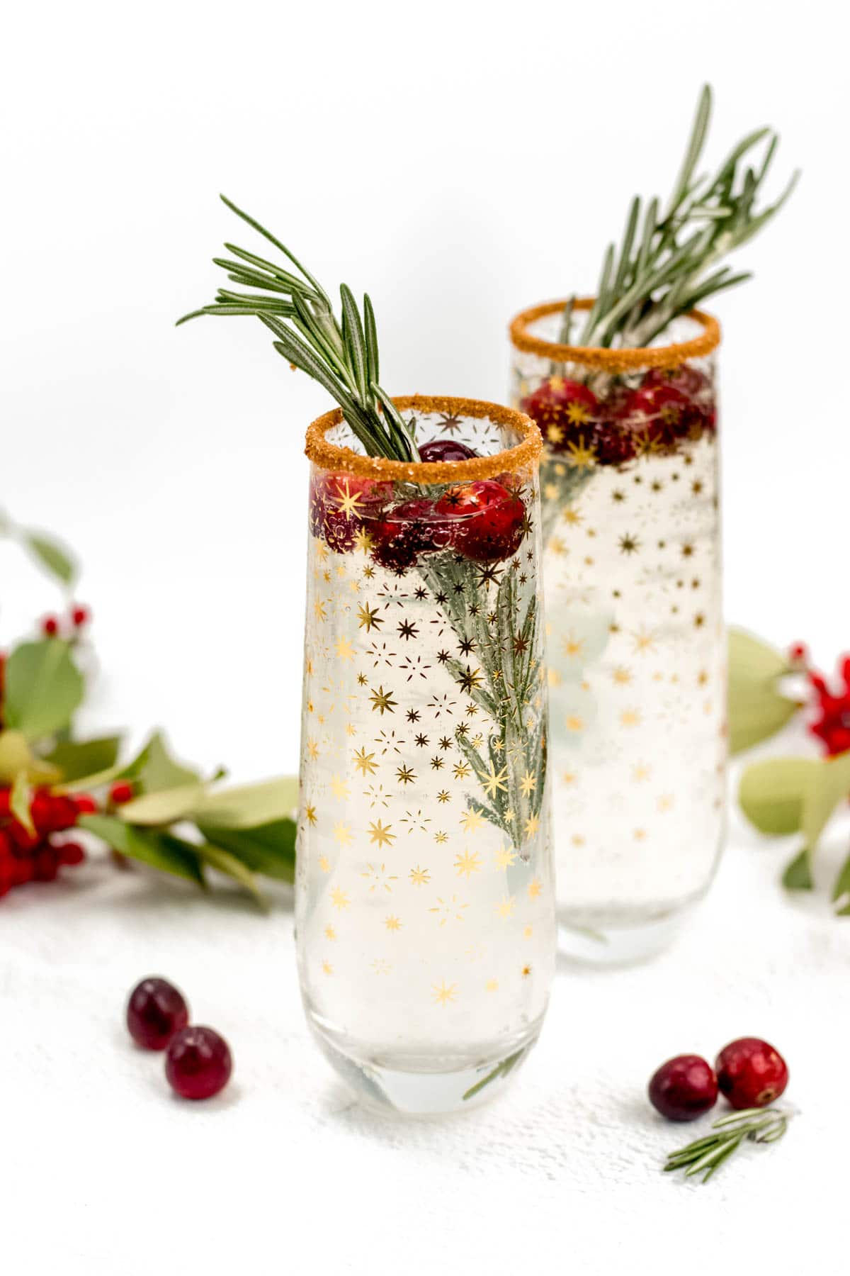 White cranberry mimosas in two stemless champagne flutes with gold stars. Cranberries and rosemary sprigs garnish alongside a cinnamon sugar rim.