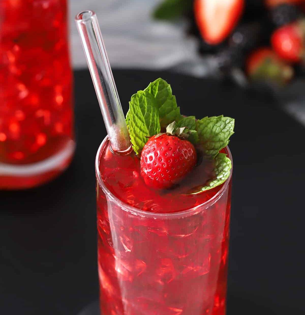 A red drink in a tall glass with a glass straw is garnished with a whole strawberry and mint sprig.