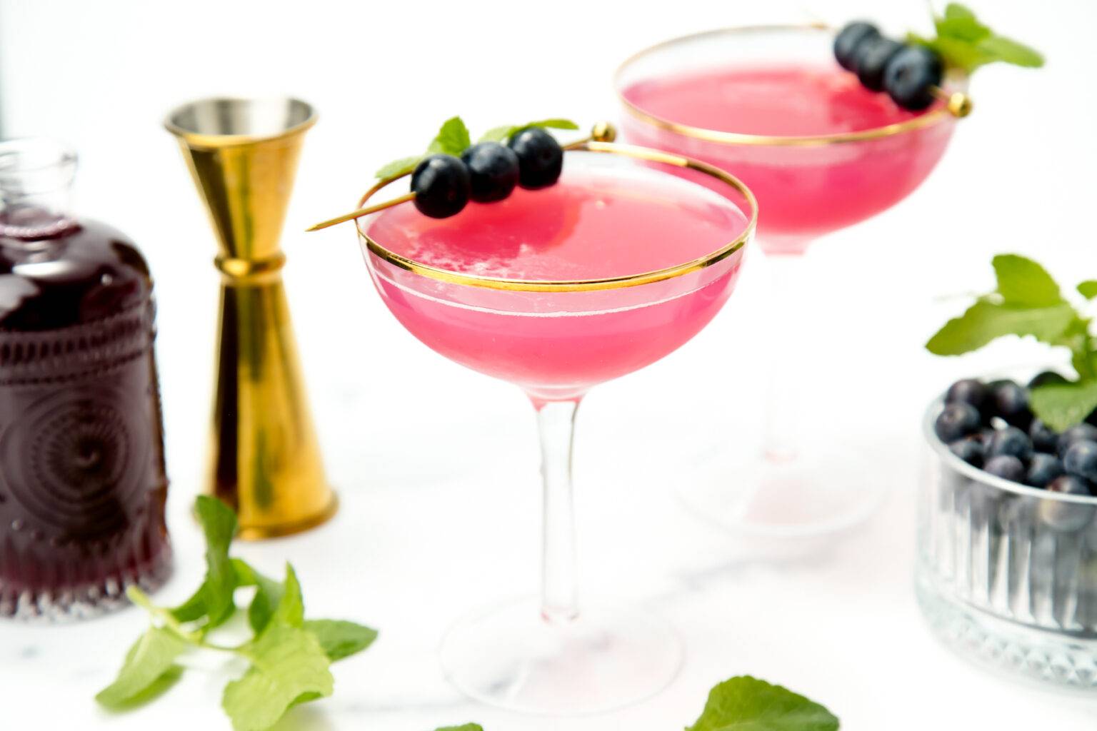 Two light purple drinks in stemmed glasses near gold bar tools.