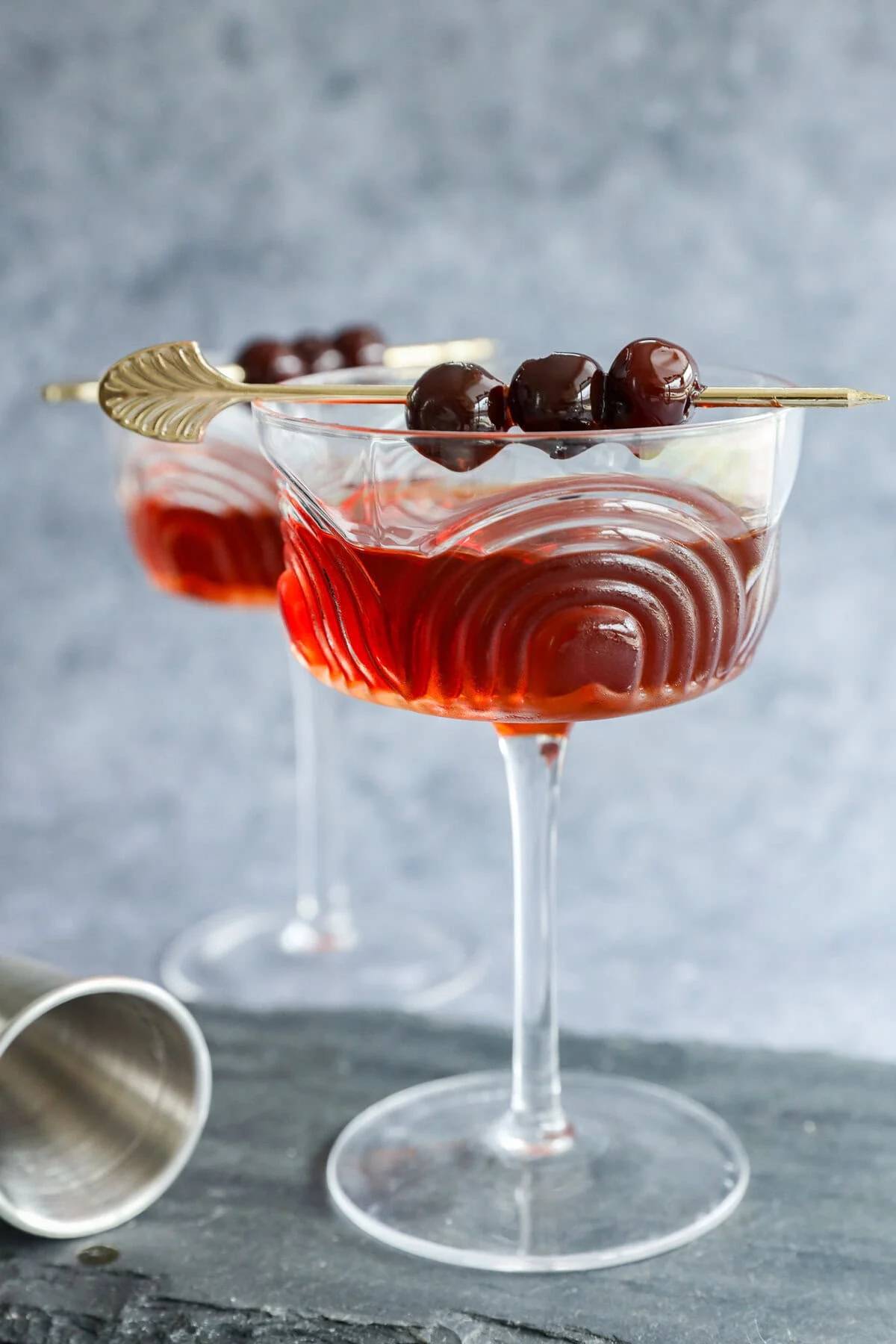 A deep amber drink in a stemmed glass topped with three cherries on a gold cocktail pick.