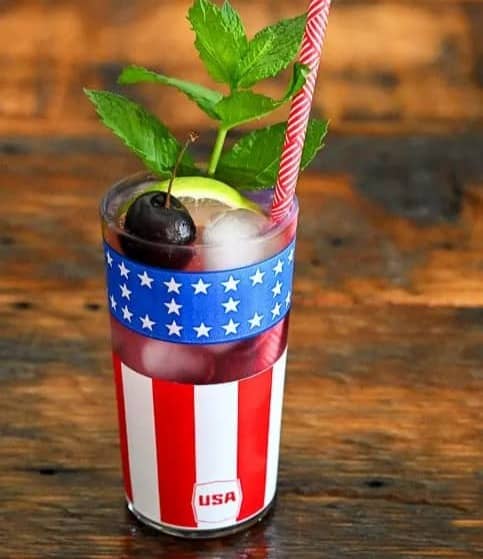A red cocktail in a glass with red and white stripes and a blue section with white stars topped with a cherry and a mint sprig.
