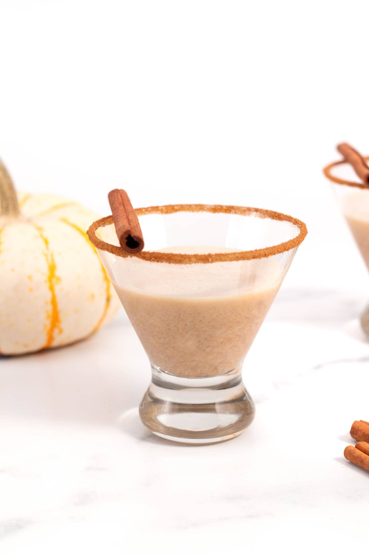 A martini in a glass rimmed with cinnamon sugar and a cinnamon stick sits near a small white pumpkin.